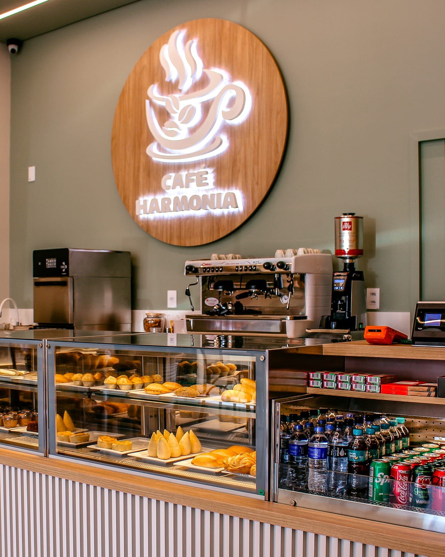 Balcão do Café Harmonia com a vitrine de salgados, a máquina de espresso, o moedor Illy e o logo iluminado na parede