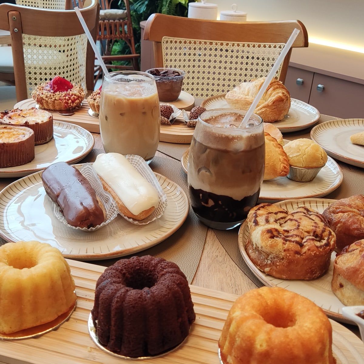 Mesa do salão com dois cafés gelados, bombas, mini bolos e folhados, e as cadeiras de palhinha ao fundo