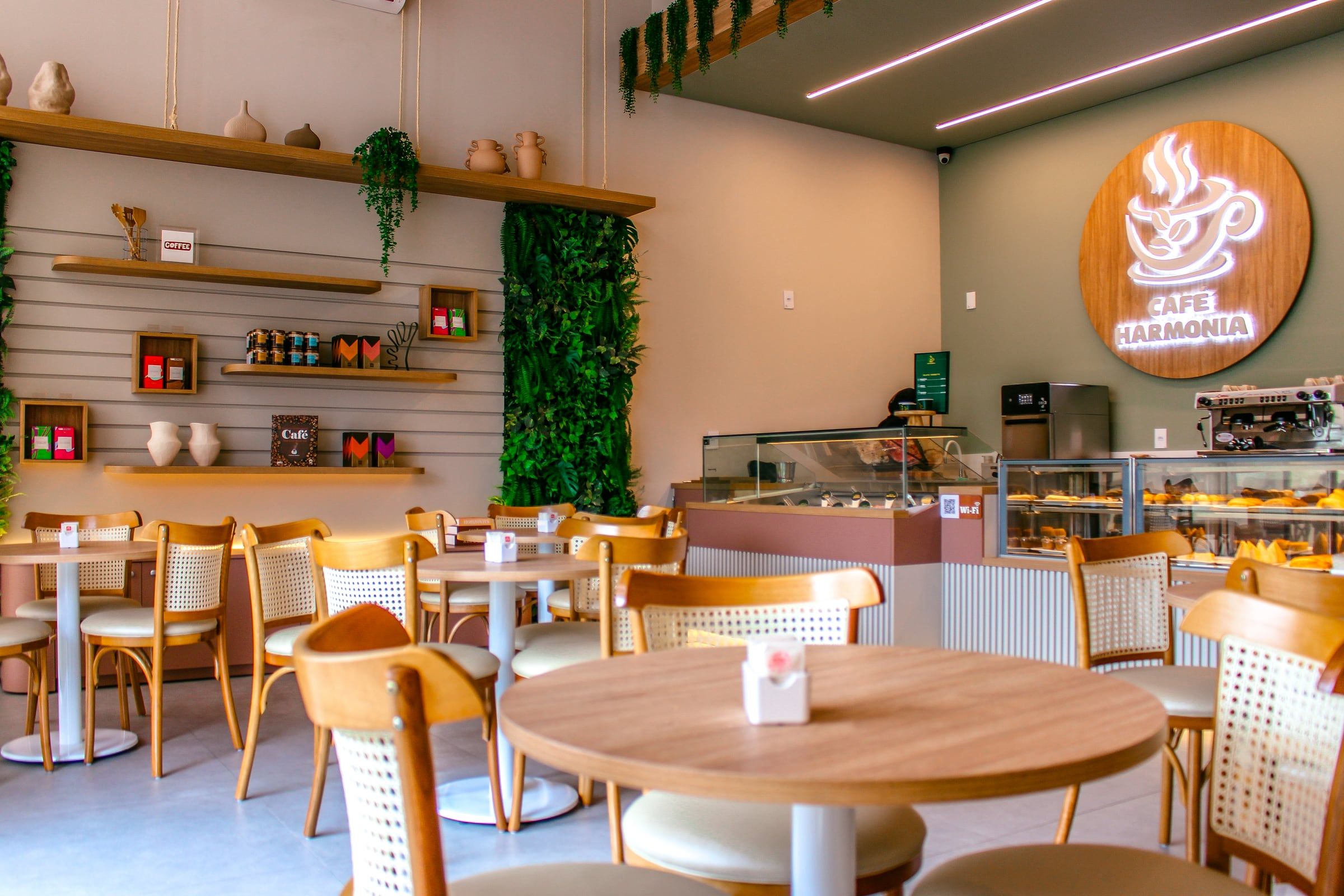 Salão do Café Harmonia com mesas redondas, cadeiras de palhinha, prateleiras com plantas e o logo aceso na parede verde