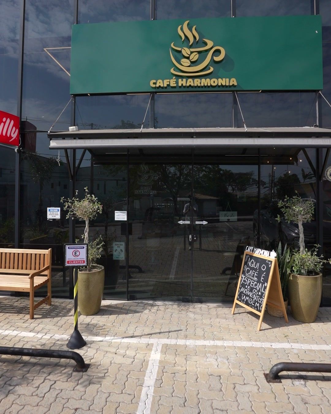 Fachada do Café Harmonia, cafeteria em Jundiaí, com letreiro verde, banco de madeira, vasos com oliveiras e vagas na frente