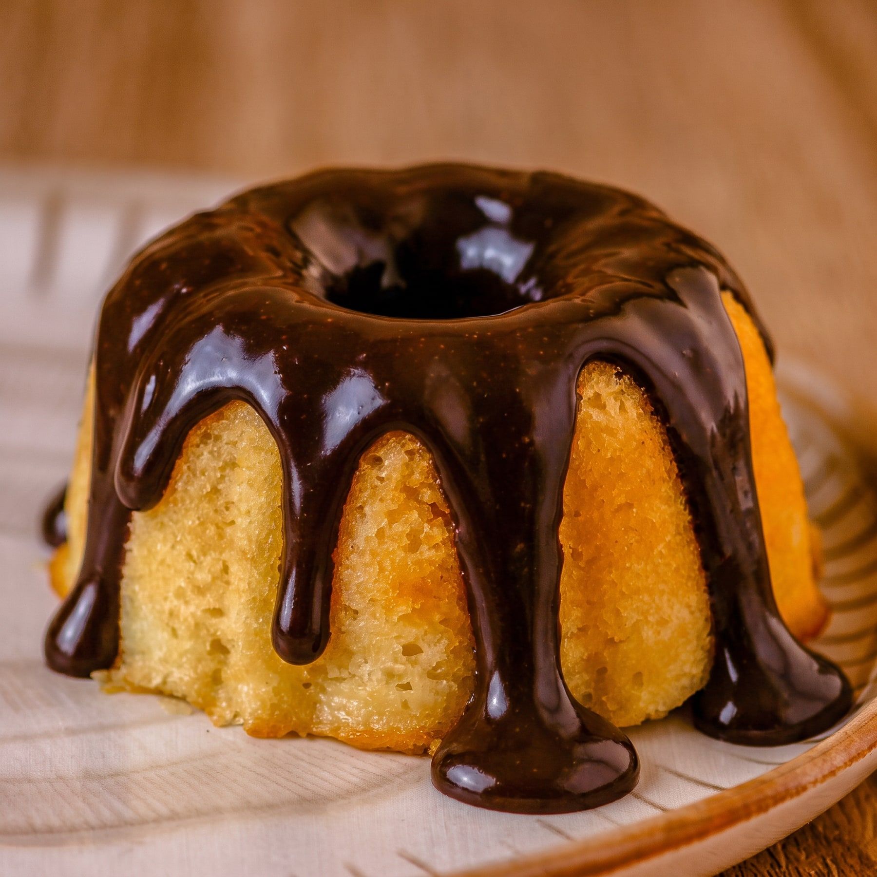 Mini bolo de laranja com calda de chocolate escorrendo pelos lados, num prato de cerâmica com a borda listrada, sobre mesa de madeira
