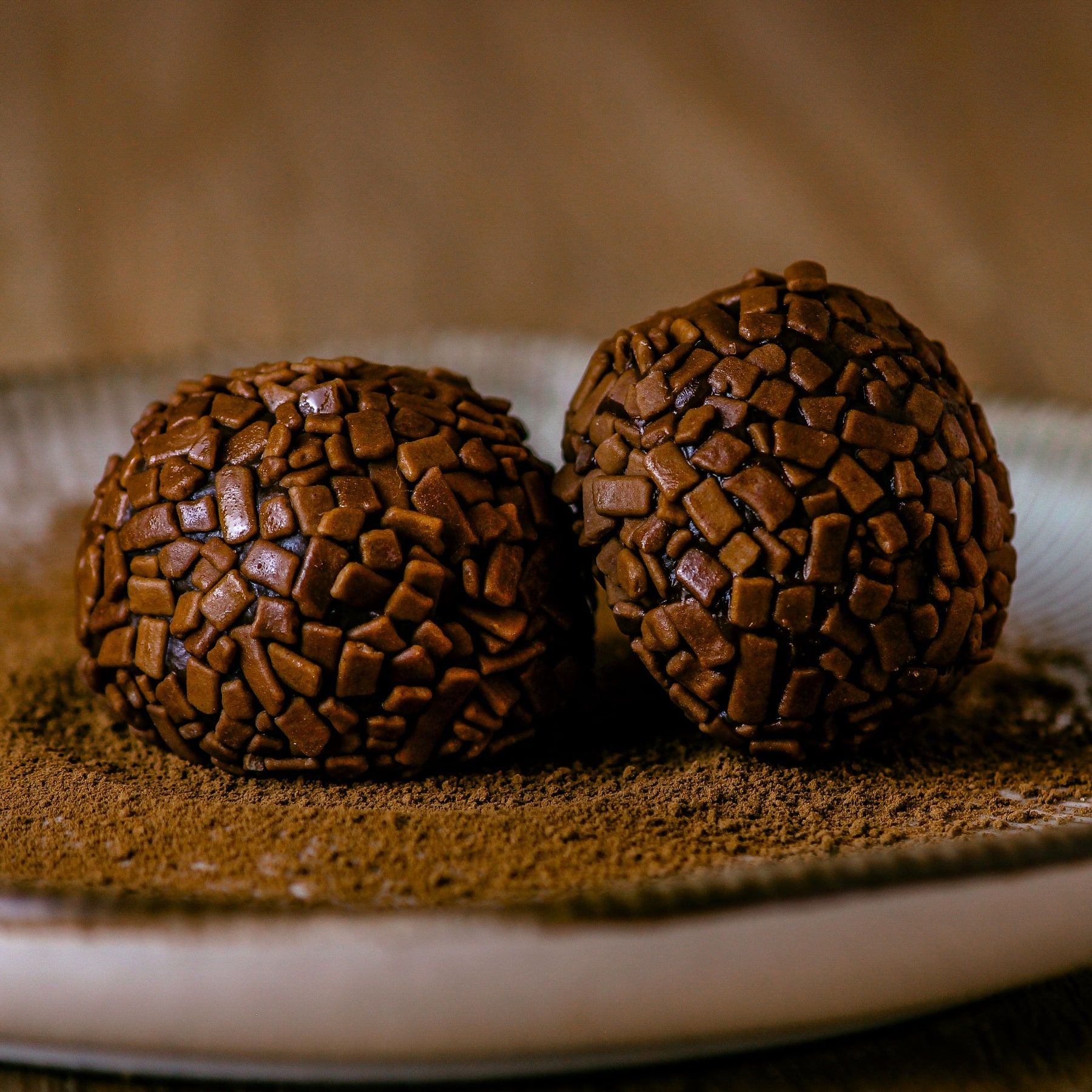 Dois brigadeiros com granulado de chocolate num prato, sobre chocolate em pó