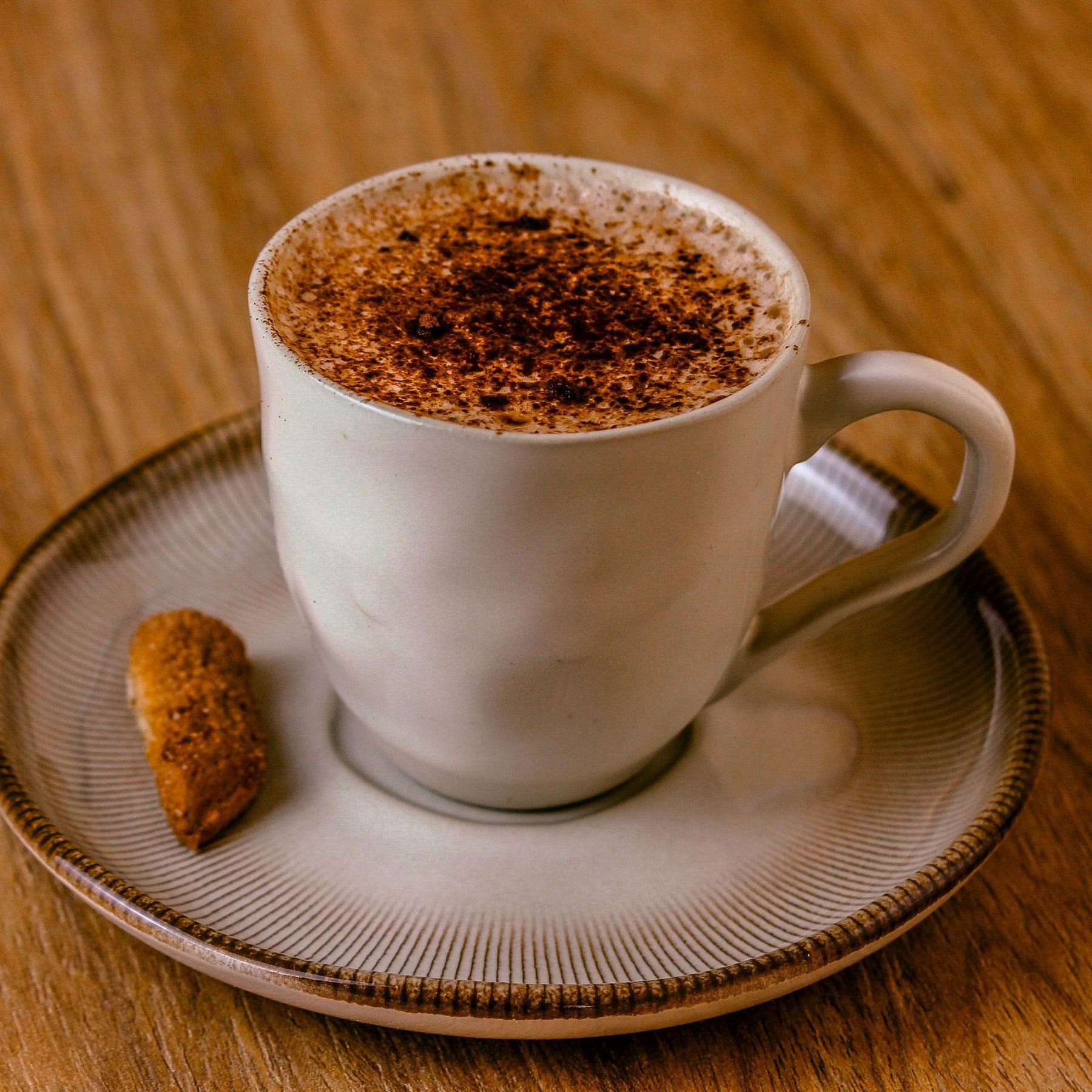 Cappuccino com cacau polvilhado na espuma, em xícara branca com um biscoito no pires, sobre mesa de madeira