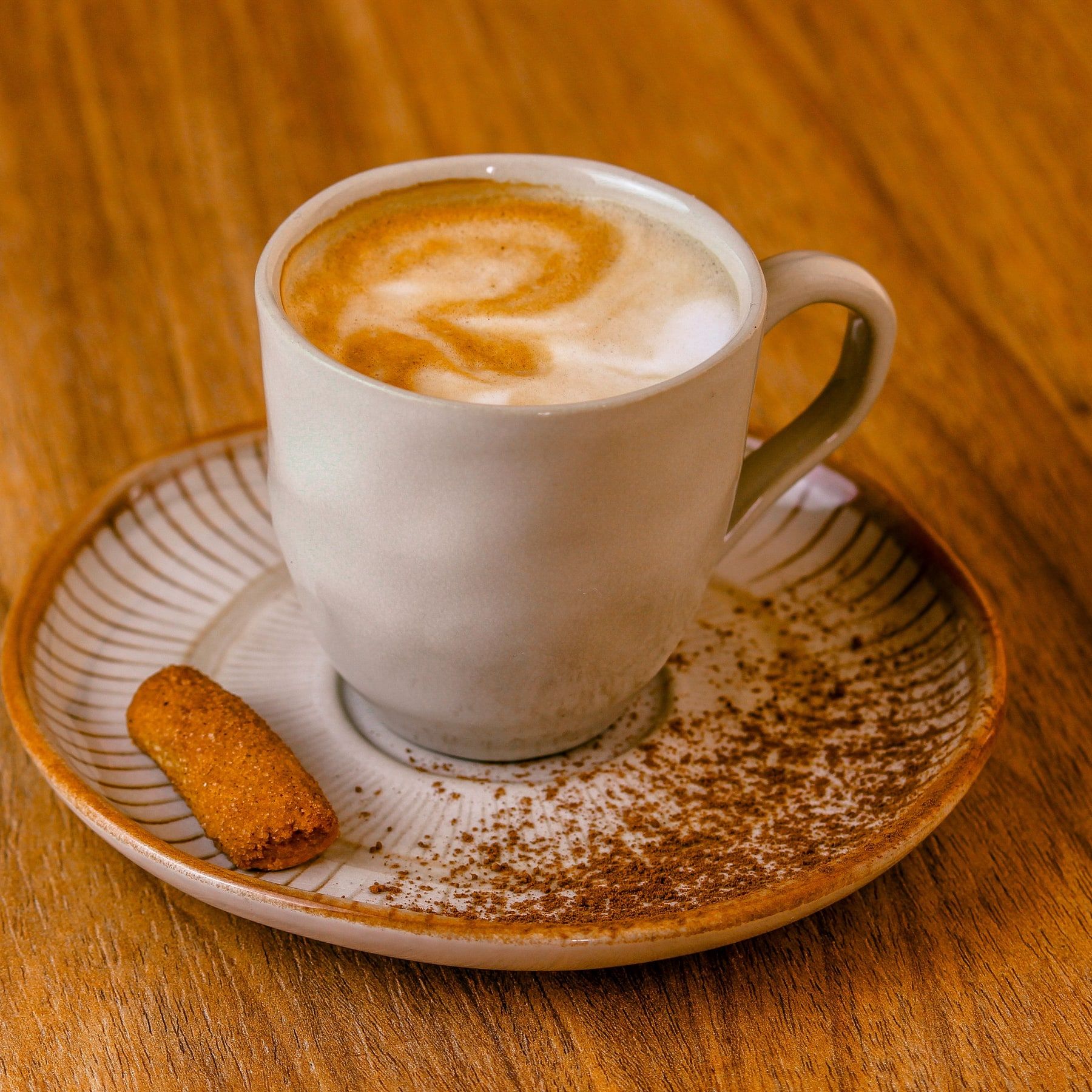 Cappuccino com desenho na espuma, em xícara branca sobre pires polvilhado de cacau, numa mesa de madeira