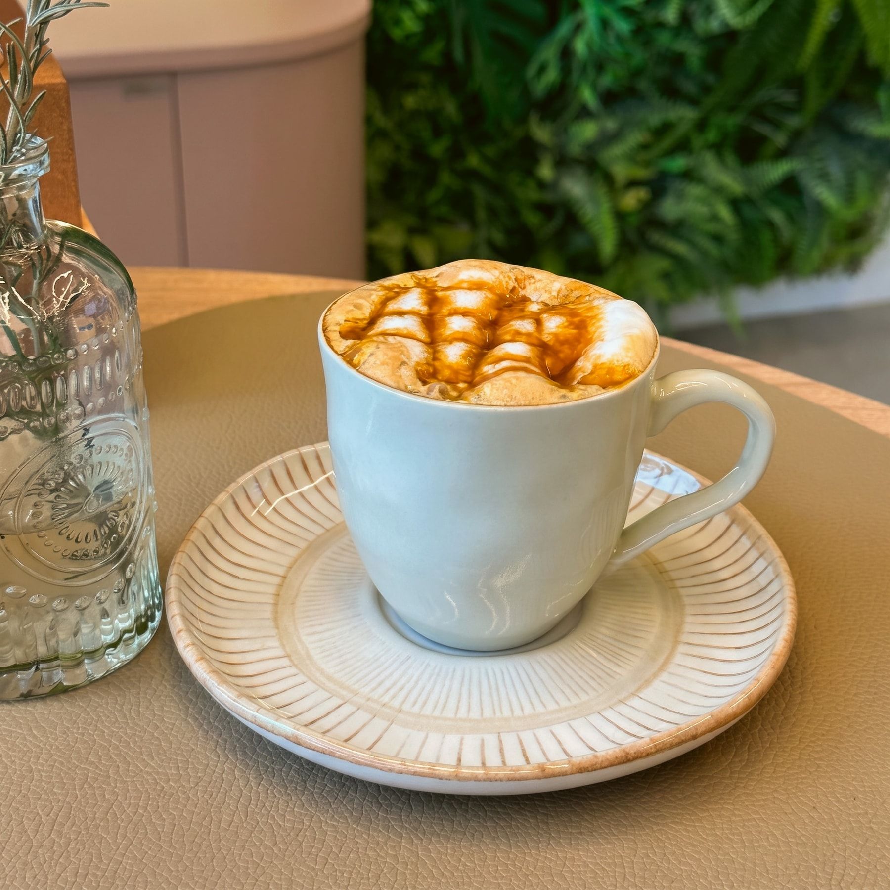Caramelo latte macchiato na xícara clara, com a espuma coberta por uma treliça de calda de caramelo, no pires listrado, com a parede de plantas ao fundo