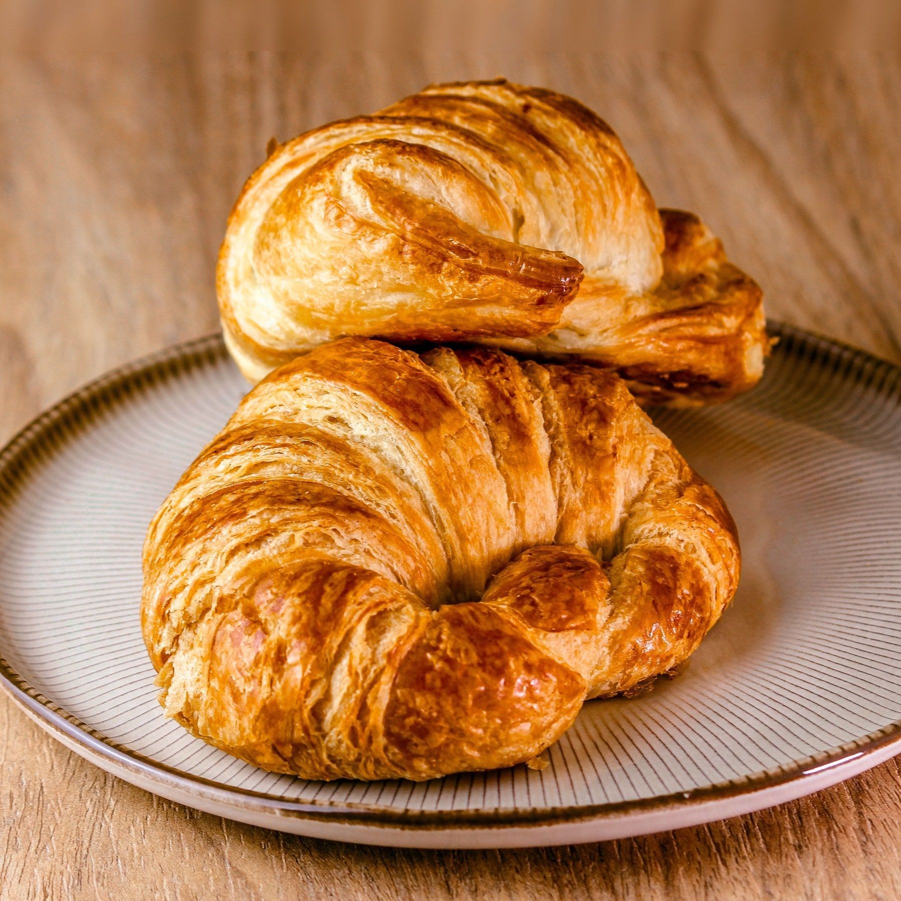 Dois croissants dourados num prato de cerâmica com a borda listrada, sobre mesa de madeira