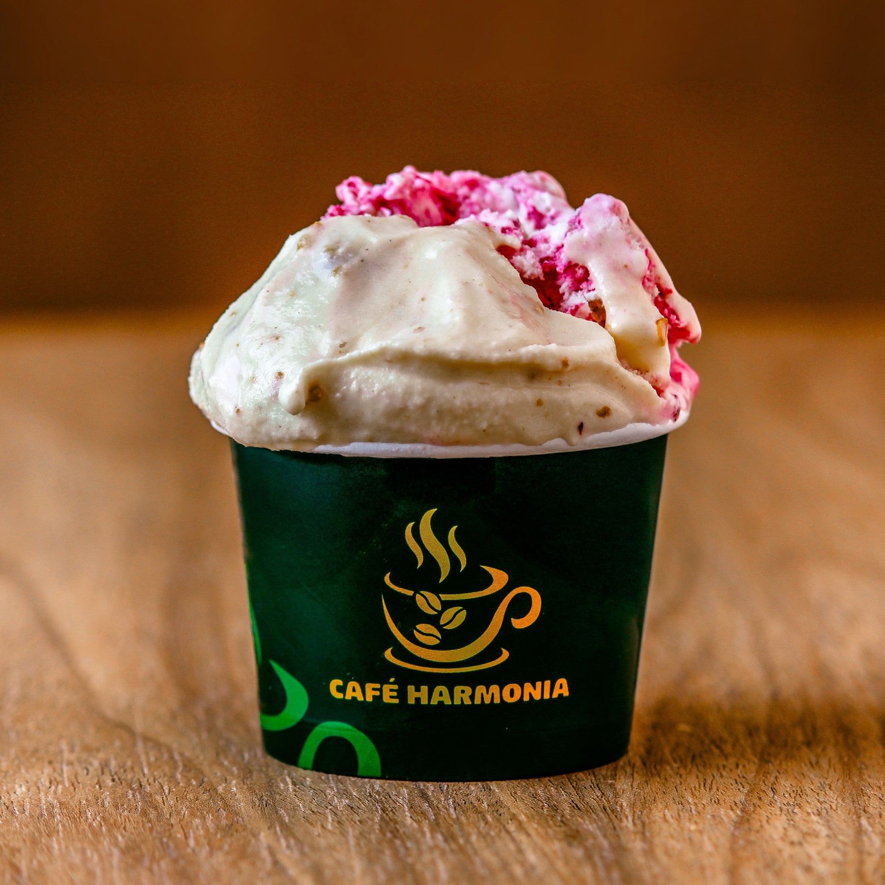 Gelato claro com pedaços cor-de-rosa no copo verde com o logo do Café Harmonia, sobre mesa de madeira