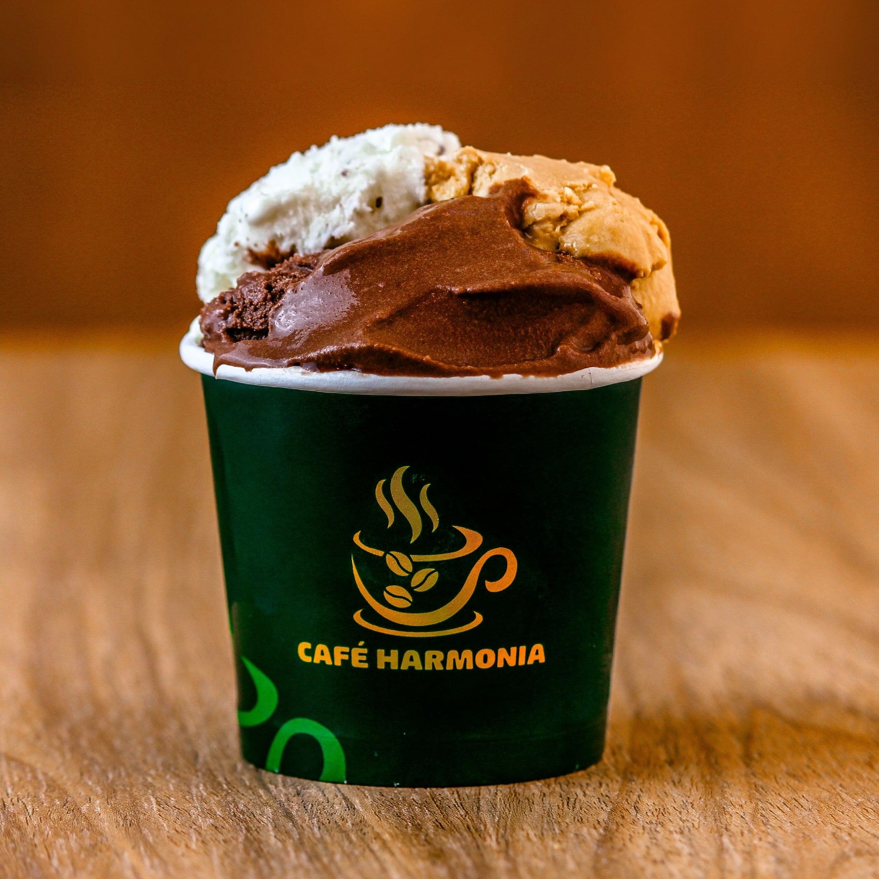Gelato de chocolate e mais dois sabores no copo verde com o logo do Café Harmonia, sobre mesa de madeira