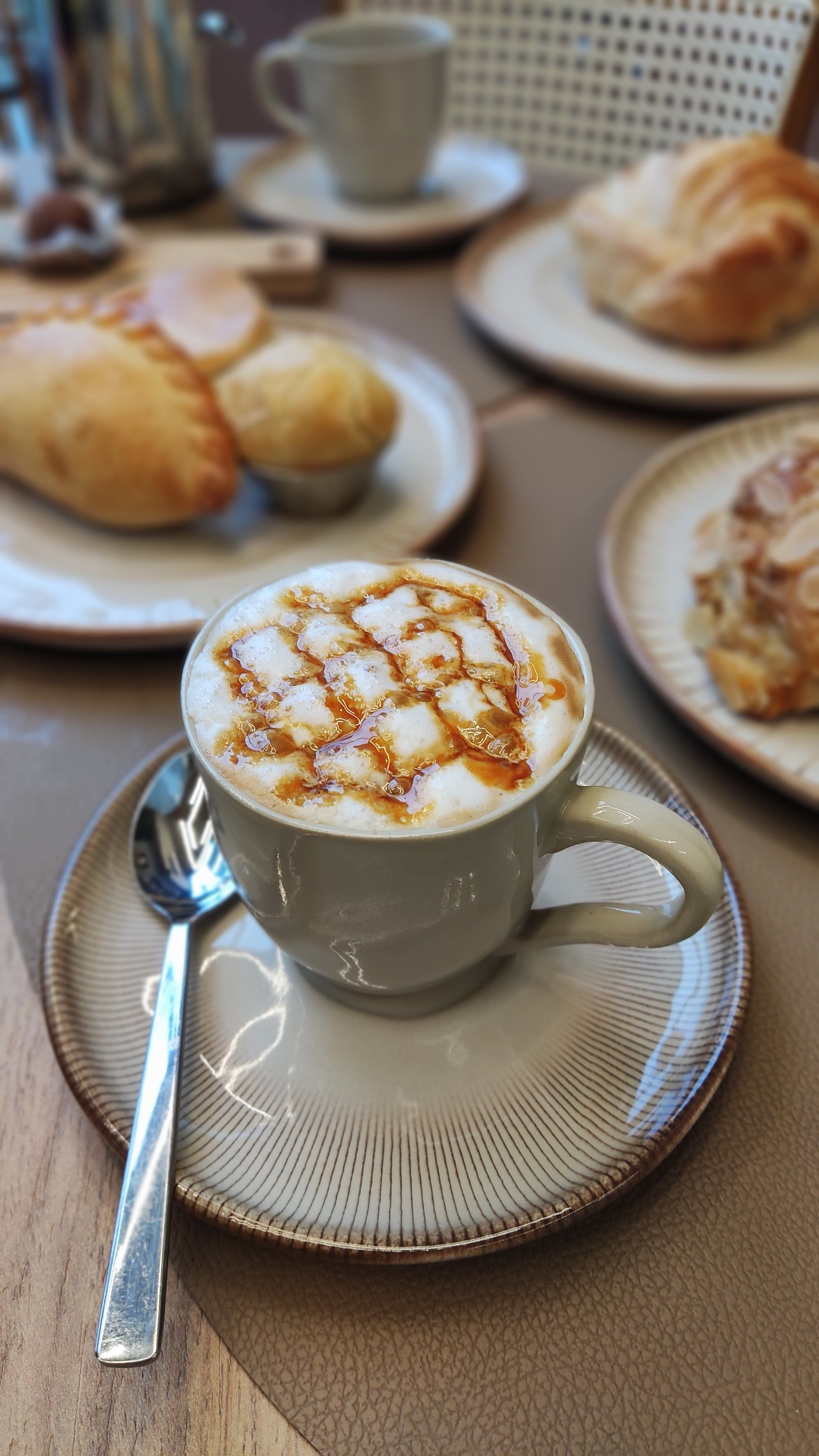Cappuccino com calda de caramelo na espuma, com salgados e croissants ao fundo na mesa do salão